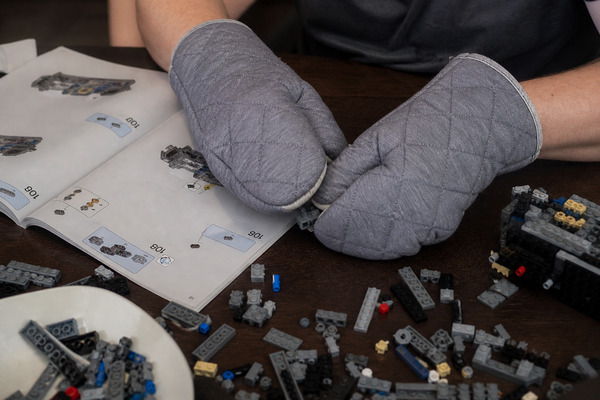 Hands in oven gloves trying to assemble small Lego pieces from the instruction manual.